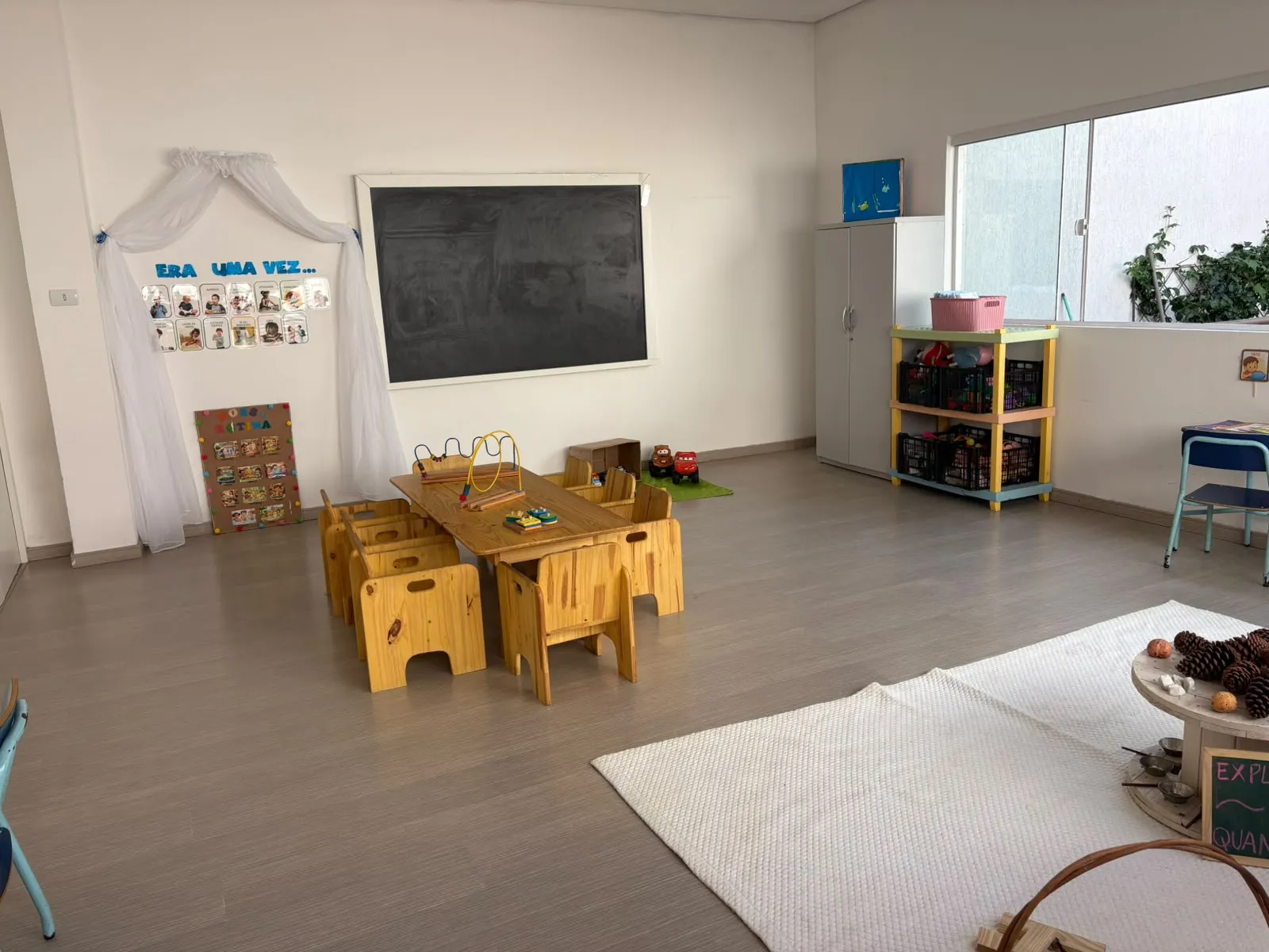 Sala de educação infantil com mesas de madeira, livros, brinquedos e iluminação natural